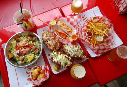 A vibrant spread of seafood dishes, fries, and drinks on a red table, featuring salads, lobster rolls, and beer.