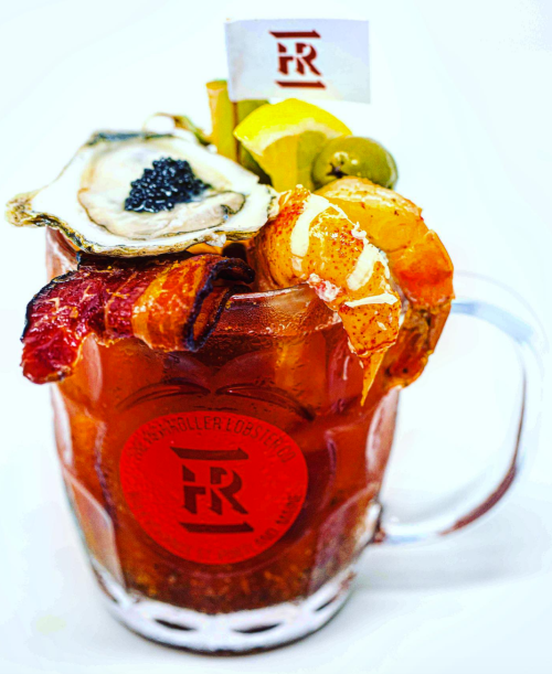 A large cocktail garnished with shrimp, bacon, an oyster, lemon, and a small flag, served in a glass mug.