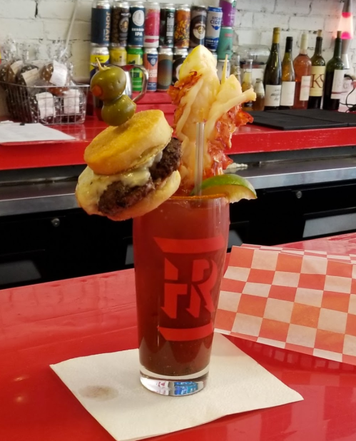 A colorful cocktail garnished with a mini burger, olives, shrimp, and bacon, served in a branded glass on a red table.