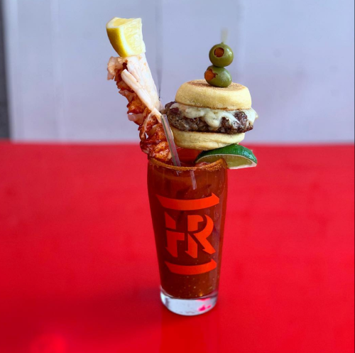 A colorful cocktail garnished with a mini burger, shrimp, olives, and lemon, served in a branded glass.
