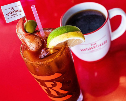 A vibrant cocktail garnished with shrimp, lime, and olive, next to a coffee mug on a bright red table.