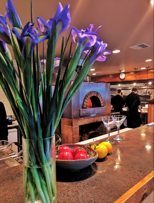 A vase of purple flowers on a countertop with a pizza oven in the background and fresh tomatoes and lemons on a plate.