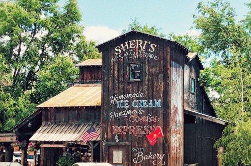 A rustic wooden shop with signs for ice cream, candies, espresso, and bakery items, surrounded by trees and blue sky.