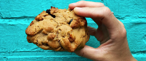 A hand holding a large, chunky cookie with nuts against a bright turquoise background.