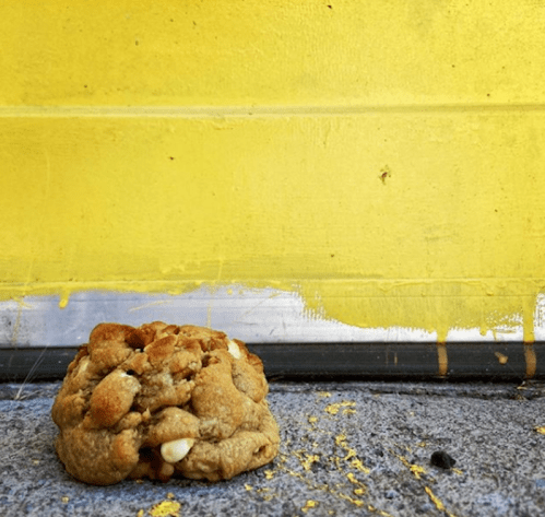 A cookie with chocolate chips sits on a textured surface next to a bright yellow wall.