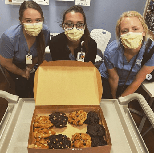Three masked individuals in blue shirts pose with a box of assorted cookies on a table.