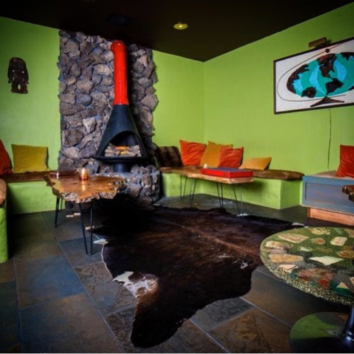 A cozy lounge with a stone fireplace, green walls, colorful cushions, and a cowhide rug on a tiled floor.