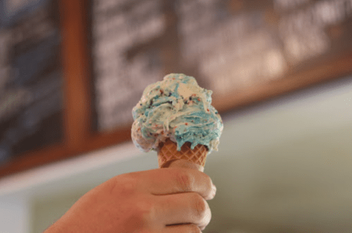 A hand holding a cone of blue and white swirled ice cream with colorful sprinkles against a blurred background.