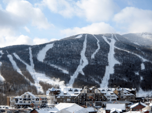 A snowy mountain with ski trails and a resort at the base, under a partly cloudy sky.