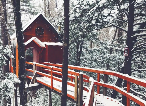 A cozy red cabin nestled in snowy trees, connected by a wooden walkway adorned with holiday lights.