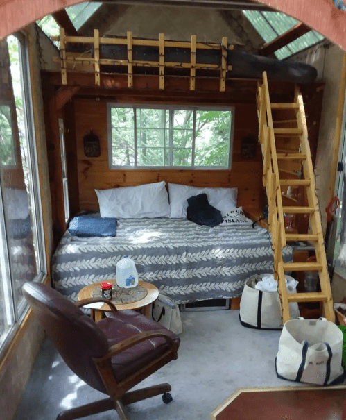 Cozy interior of a small cabin with a loft bed, wooden ladder, and a seating area by a window.