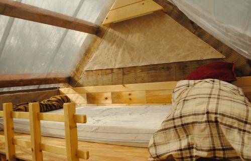 Cozy, rustic interior of a small cabin with a wooden bed, plaid blankets, and a sloped ceiling.