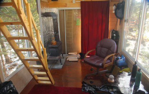 Cozy interior of a cabin with a wooden staircase, a red curtain, a chair, and a wood stove, surrounded by nature.