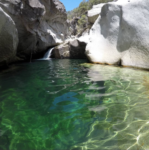 A serene natural pool surrounded by large rocks and a small waterfall, with clear green water reflecting sunlight.