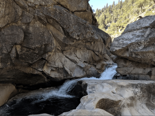 A rocky riverbed with flowing water surrounded by steep cliffs and lush greenery in a natural landscape.