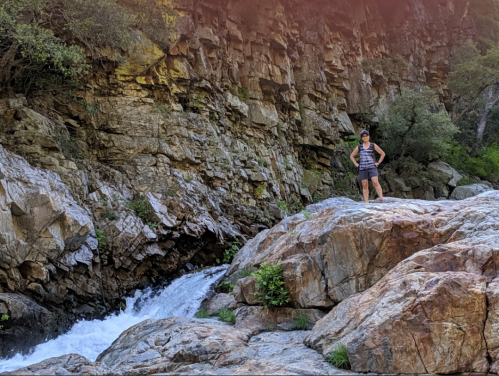 A person stands on a rocky outcrop near a flowing stream, surrounded by lush greenery and steep rock formations.