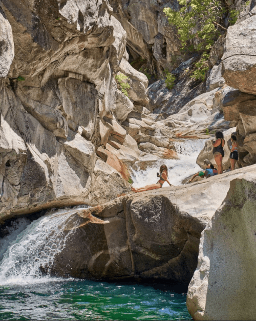 A group of people relaxes by a rocky riverbank with cascading water and lush greenery in a sunny outdoor setting.