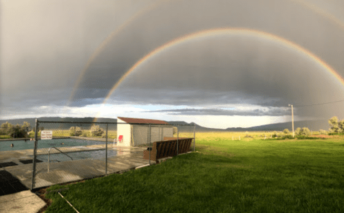 A serene landscape featuring a swimming pool, green grass, and a double rainbow against a cloudy sky.