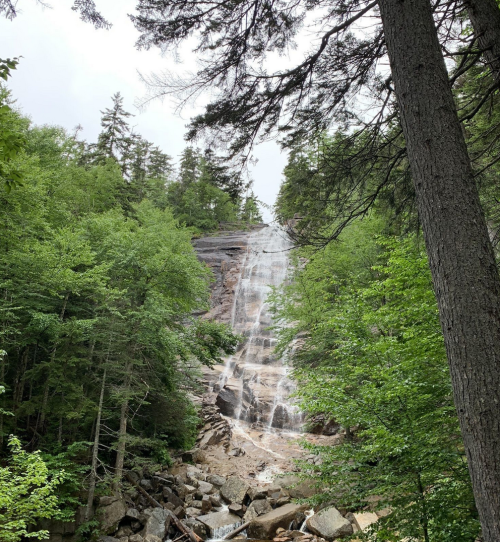A waterfall cascades down rocky terrain, surrounded by lush green trees and a cloudy sky.