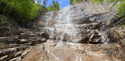 A cascading waterfall flows down a rocky cliff surrounded by lush green trees under a clear blue sky.