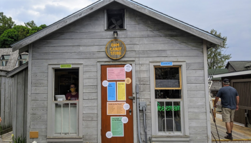 A small wooden candy store with colorful signs, featuring a woman at the counter and a man walking by outside.
