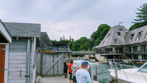 People stand by a wooden dock near a candy store, with boats and buildings along a flowing river.