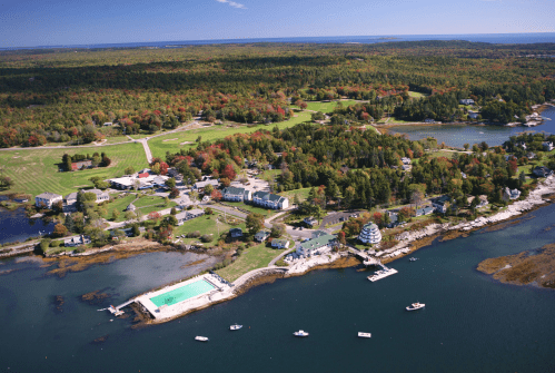 Aerial view of a coastal area with lush greenery, a pool, and boats along the shoreline under a clear blue sky.