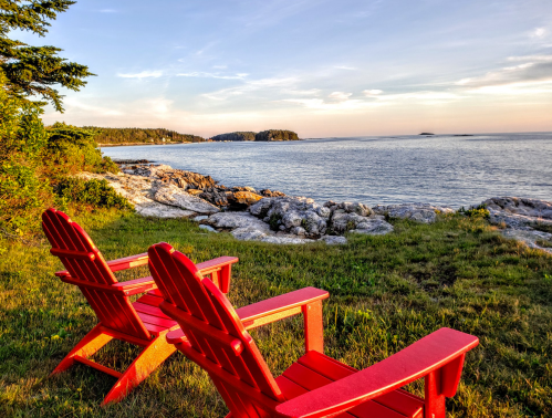 Two red Adirondack chairs sit on a grassy shore, overlooking a calm sea and rocky coastline at sunset.