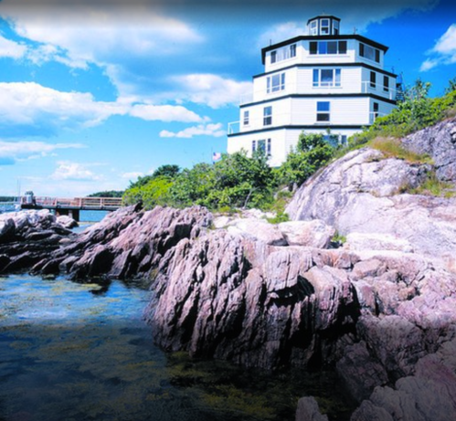A coastal scene featuring a white house on a rocky shore under a bright blue sky with scattered clouds.