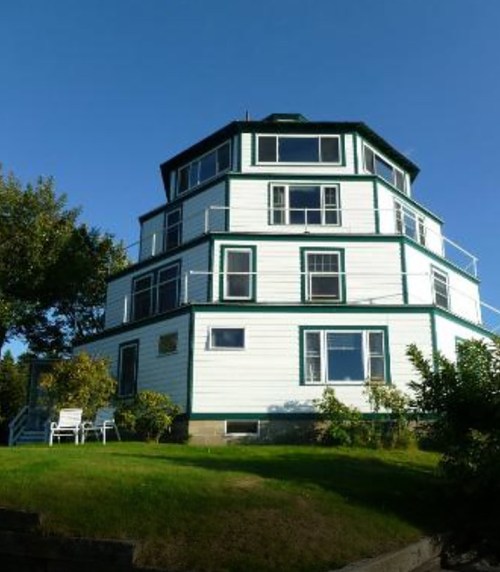 A unique, multi-level octagonal house with large windows and green trim, surrounded by grass and trees.