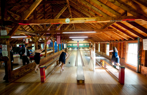 A rustic bowling alley with wooden beams, featuring four people in dresses bowling. Colorful lights add a festive touch.