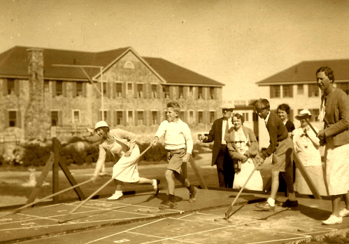 A vintage photo of children and adults playing a game outdoors, with a stone building in the background.
