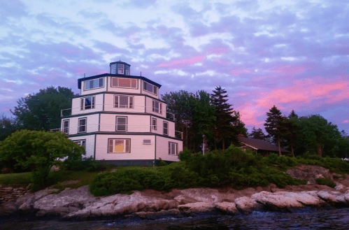 A unique octagonal house by the water, surrounded by trees, with a colorful sunset sky in the background.