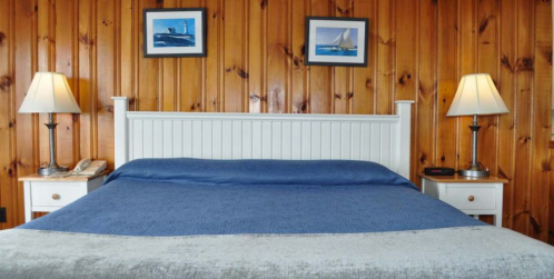 Cozy bedroom with a blue bedspread, wooden walls, and framed nautical art, featuring bedside lamps and a phone.