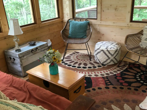 Cozy interior of a cabin with wooden walls, a sofa, chairs, a coffee table, and a colorful rug. Flowers in a vase.
