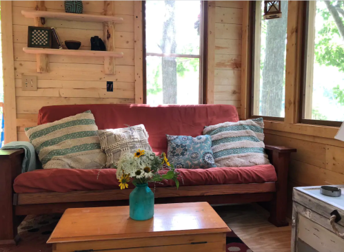 Cozy interior with a red couch, decorative pillows, a wooden coffee table, and a vase of flowers in a sunlit room.