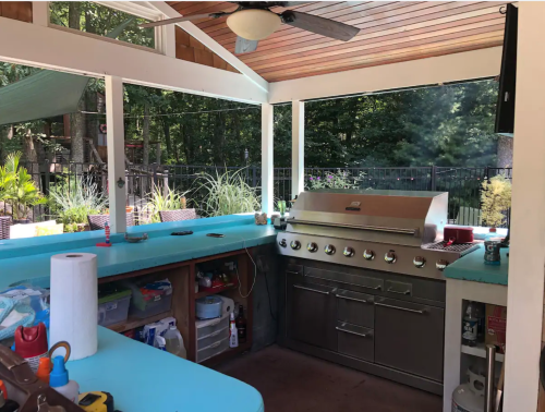 A modern outdoor kitchen with a grill, turquoise countertops, and a ceiling fan, surrounded by greenery.