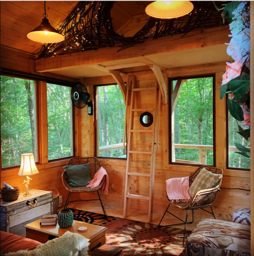 Cozy wooden cabin interior with a ladder, seating area, and large windows overlooking a forest. Warm lighting adds charm.
