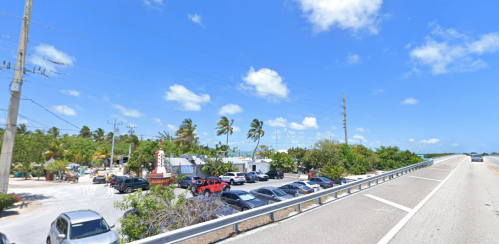 A sunny roadside view with parked cars, palm trees, and a sign for a nearby establishment.