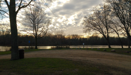 A serene lakeside view at sunset, with trees, clouds, and a dirt path leading to the water.