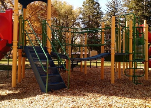 Colorful playground equipment with slides, climbing structures, and a bridge, surrounded by trees and wood chips.