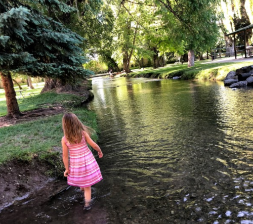 A young girl in a striped dress wades through a shallow stream in a lush, green park.