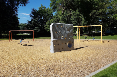 A playground featuring a climbing wall, swings, and a bench, surrounded by wood chips and trees under a clear blue sky.
