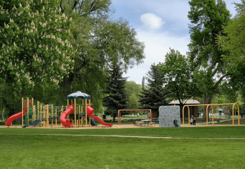 A colorful playground with slides, climbing structures, and swings, surrounded by trees and green grass.