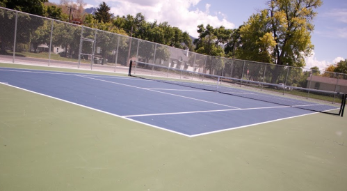 A blue tennis court with a net, surrounded by a fence and trees under a partly cloudy sky.