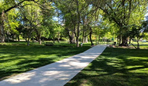 A paved path winds through a lush green park with trees and benches, inviting visitors to enjoy the outdoors.