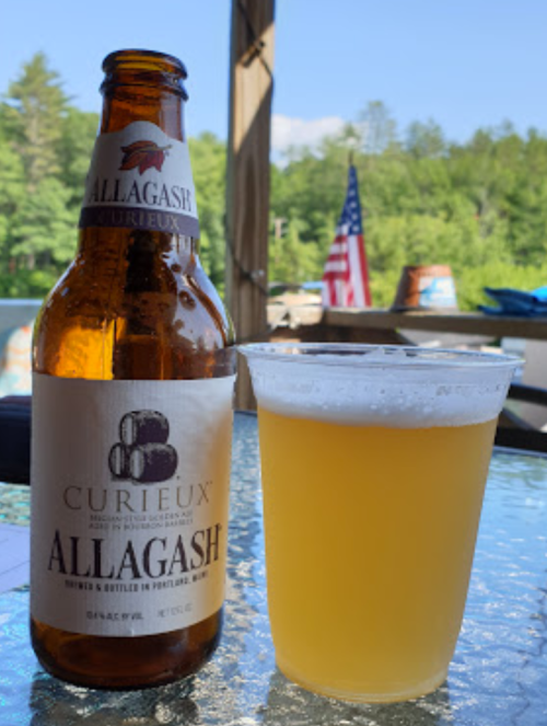 A bottle of Allagash Curieux beer next to a clear cup of beer, with a scenic outdoor background and an American flag.