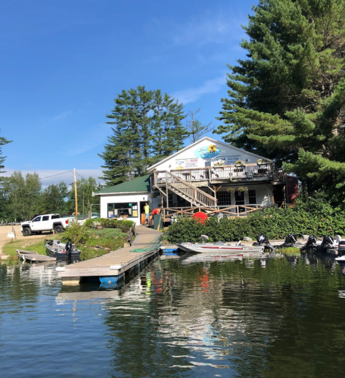 A lakeside building with a deck, surrounded by trees and boats, reflecting in the water under a clear blue sky.
