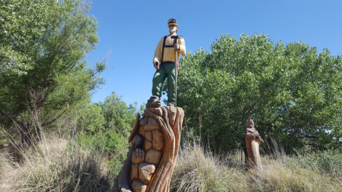 A wooden statue of a man in work clothes stands atop a carved base, surrounded by greenery and blue sky.