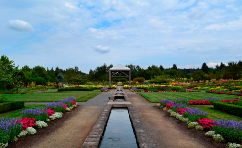 A vibrant garden with colorful flowers, a central water feature, and a gazebo under a cloudy sky.